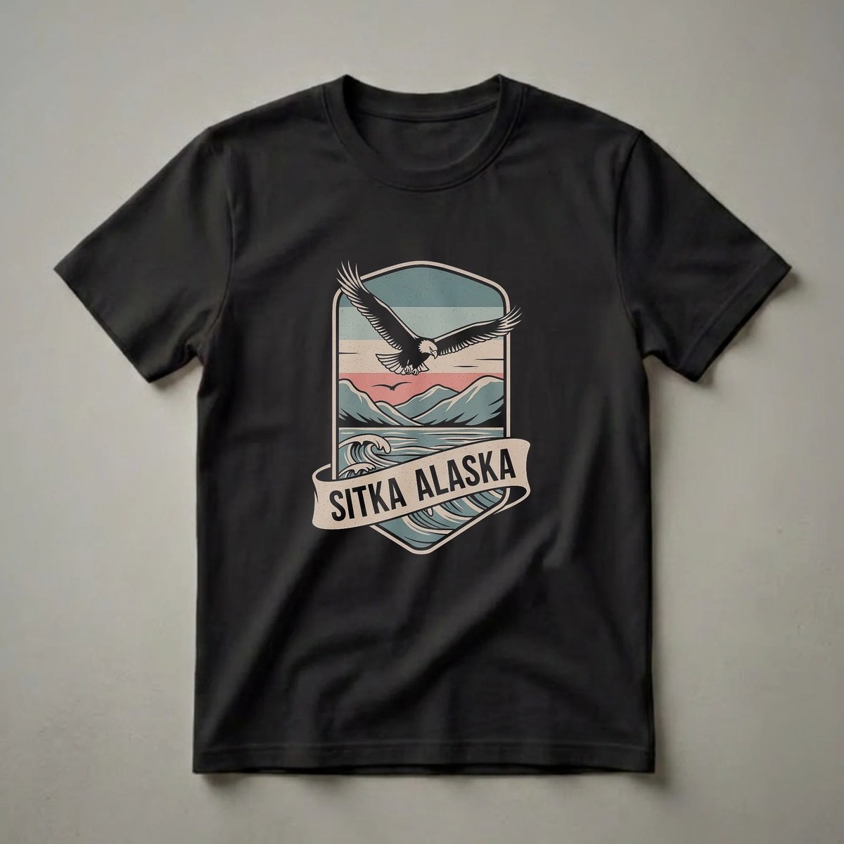 Retro graphic t-shirt featuring a black eagle in flight above stylized mountains and waves, with 'Sitka Alaska' text on a banner. The design uses a muted color palette of teal, cream, and red.