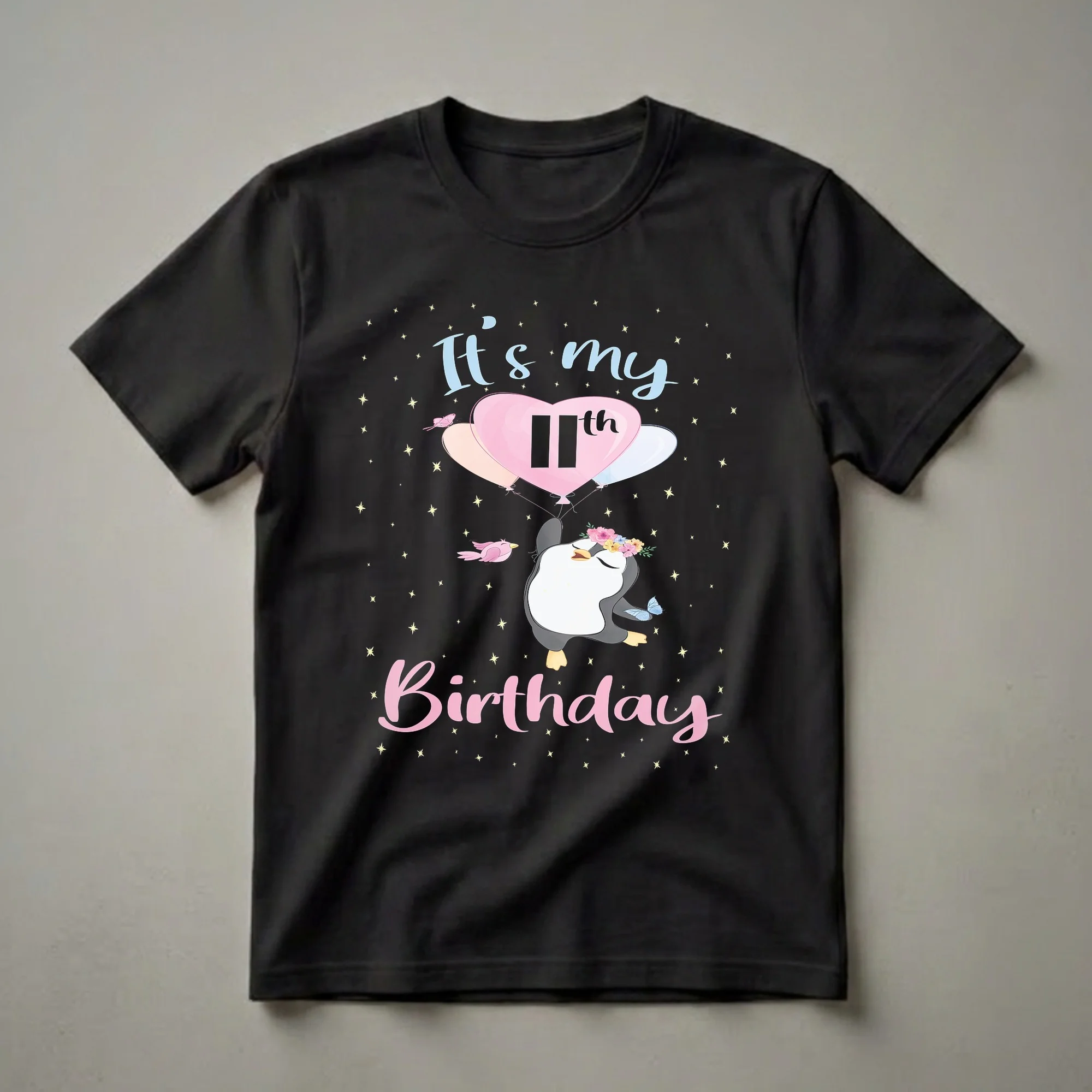 Black t-shirt featuring a cartoon penguin wearing a flower crown, holding balloons with '11th' on a heart balloon, surrounded by stars, birds, and butterflies, with 'It's my Birthday' text.