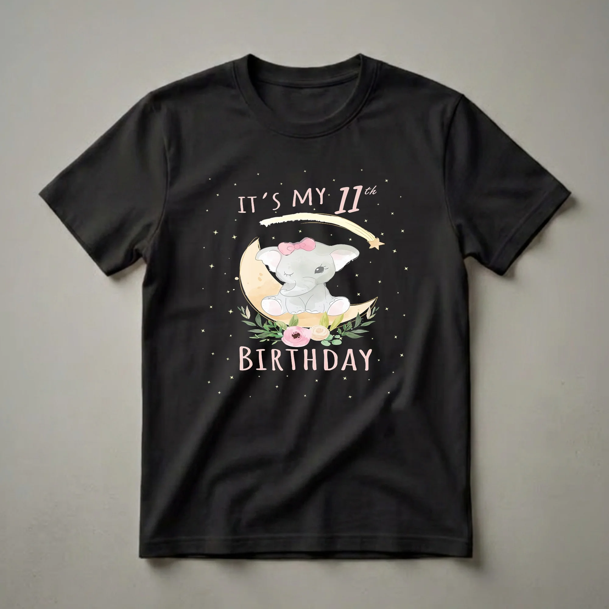Black t-shirt featuring a watercolor illustration of a baby elephant with a pink bow sitting on a crescent moon, surrounded by stars and flowers, with the text 'IT'S MY 11th BIRTHDAY' in pink.