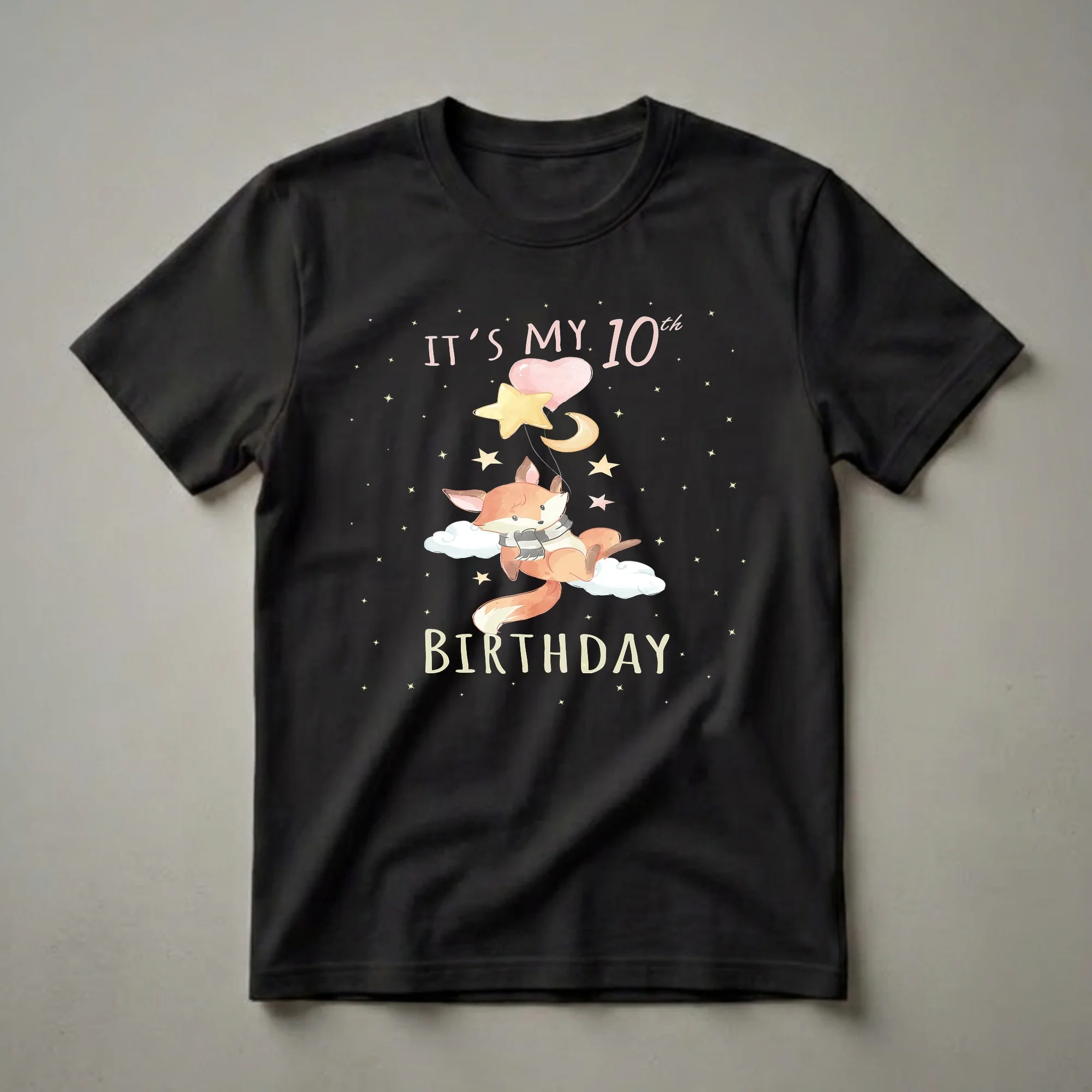 Black t-shirt featuring a watercolor illustration of a fox wearing a striped scarf, floating on clouds, holding balloons and celestial shapes against a starry night sky, with text 'IT'S MY 10th BIRTHDAY'.
