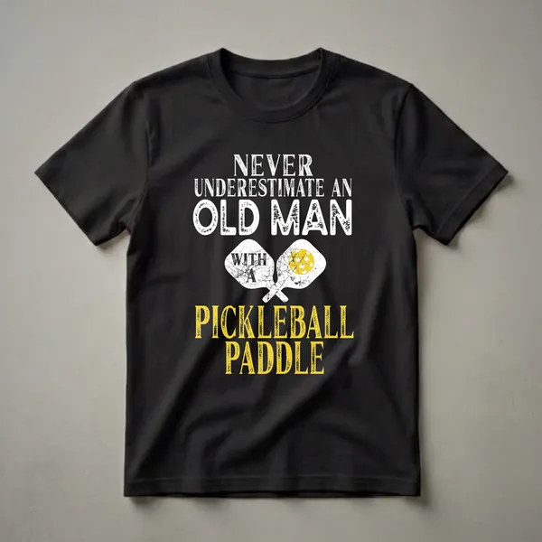 Black t-shirt with white and yellow distressed graphic text that reads 'Never Underestimate An Old Man With A Pickleball Paddle', featuring two crossed pickleball paddles.