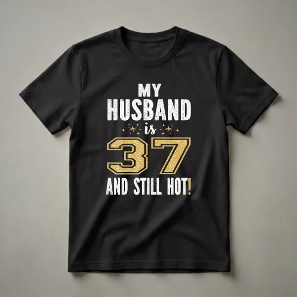Black t-shirt with white distressed text reading 'MY HUSBAND IS 37 AND STILL HOT!' and a large gold textured number 37 with sparkling accents.
