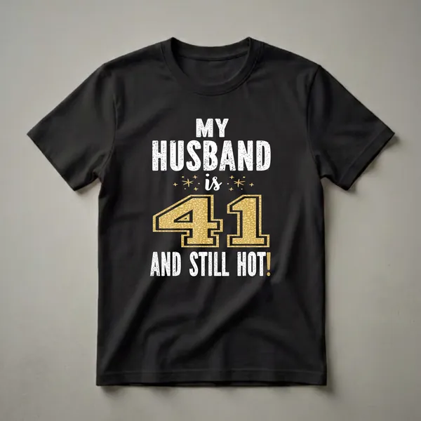 Black t-shirt with white distressed text reading 'MY HUSBAND is 41 AND STILL HOT!' with a large gold textured number 41 and small gold starbursts.