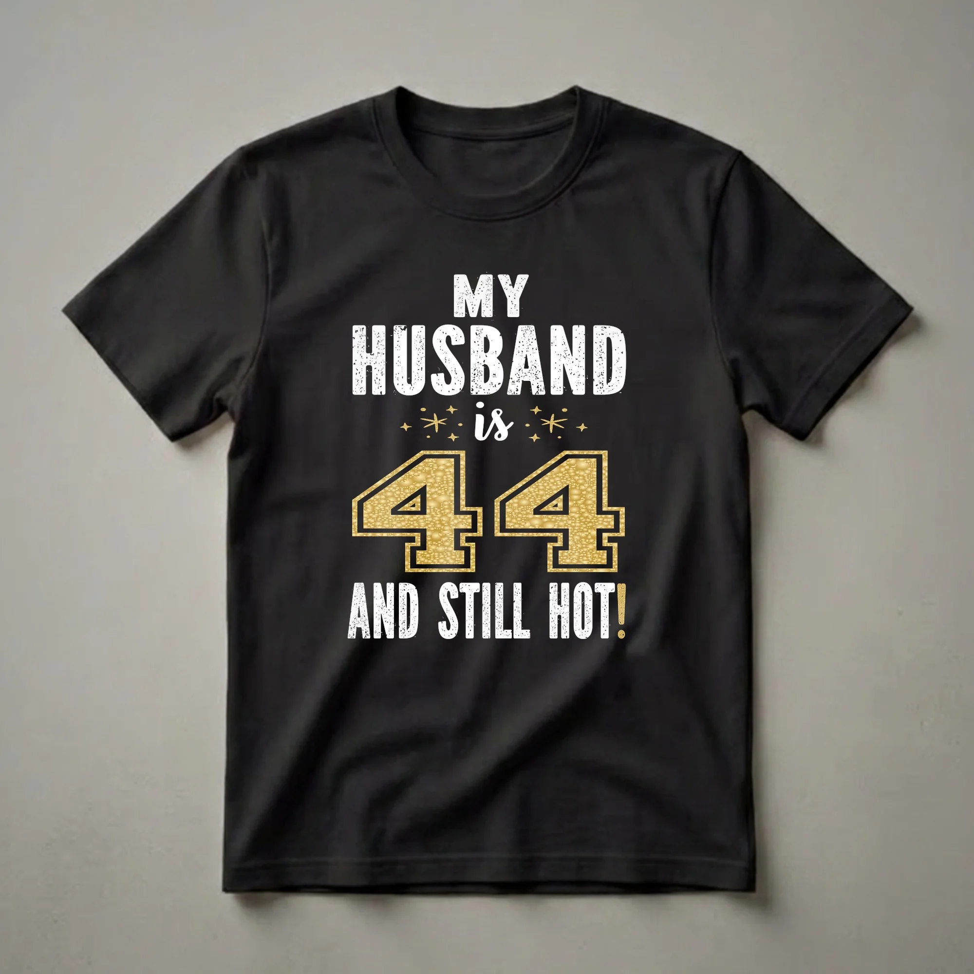 Black t-shirt with white distressed text reading "MY HUSBAND IS" and "AND STILL HOT!", with a large gold glitter "44" in the center and small gold star accents.