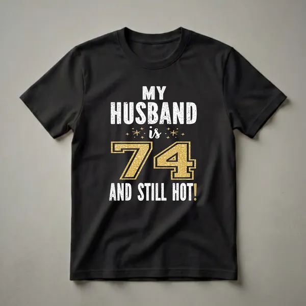 Black t-shirt with white distressed text reading 'MY HUSBAND is 74 AND STILL HOT!' with gold glitter numbers and accents.