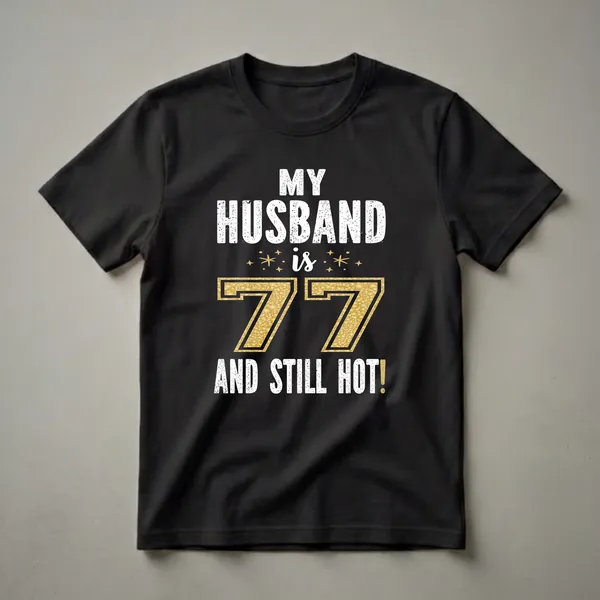 Black t-shirt with white distressed text reading 'MY HUSBAND IS 77 AND STILL HOT!' and a large gold textured number 77.