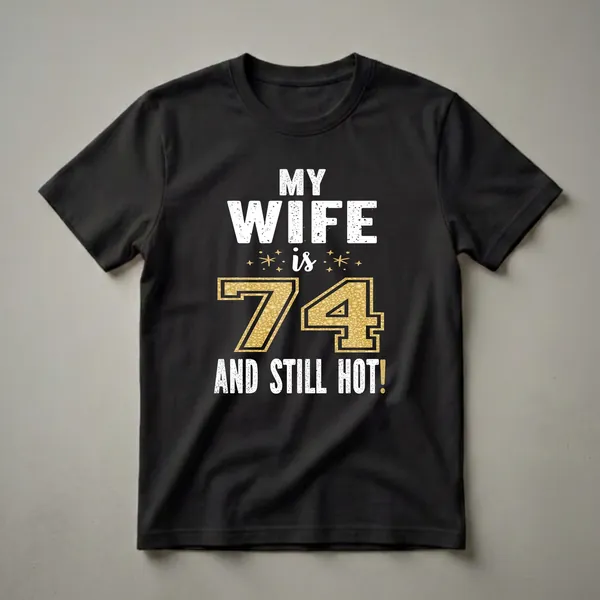 Black t-shirt with white distressed text reading 'MY WIFE is 74 AND STILL HOT!' with a large gold textured number 74 and gold star accents.