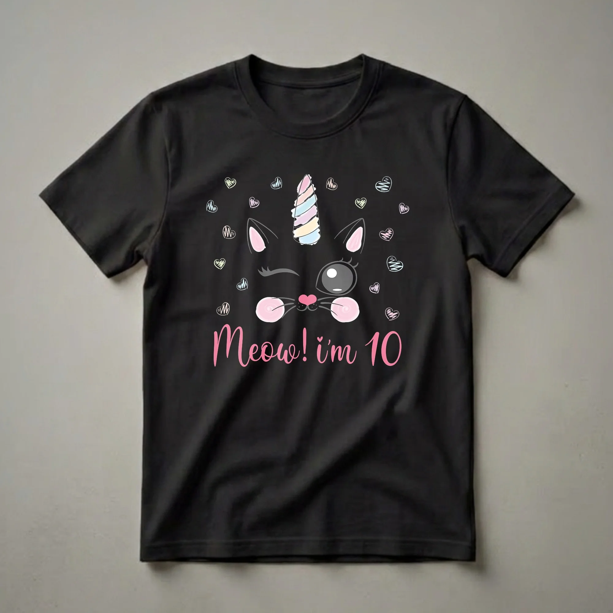 Black t-shirt graphic featuring a winking cat face with a pastel unicorn horn, pink cheeks, and the text "Meow! I'm 10", surrounded by small, colorful sketched hearts.