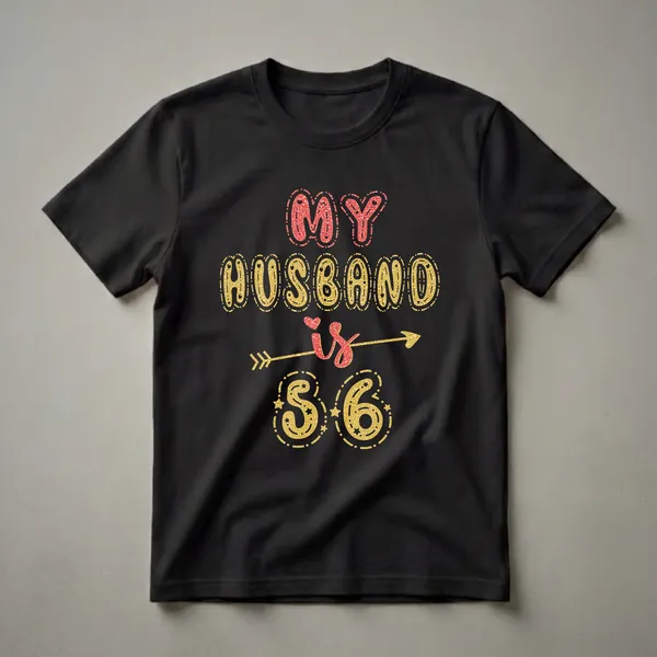 Black t-shirt with 'MY HUSBAND is 56' text graphic in red and gold glitter effect, with an arrow and stars.