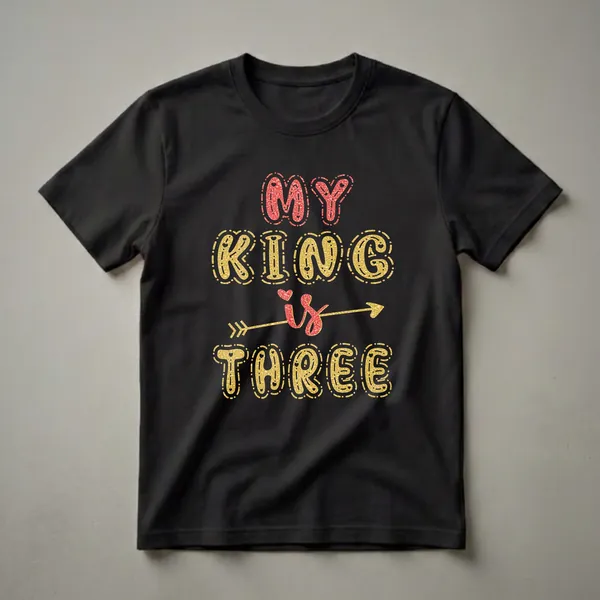 A black t-shirt with the text "MY KING is THREE" in glittery red and gold lettering, with a golden arrow through the word "is".