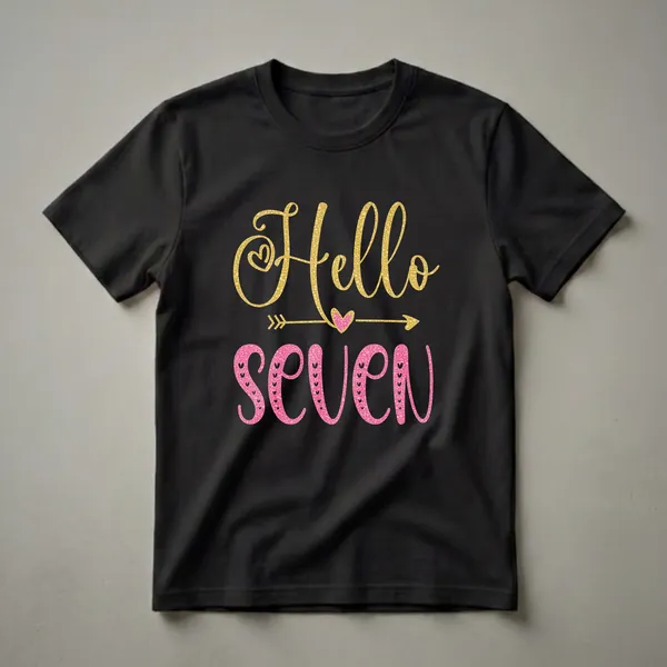 Black t-shirt with 'Hello Seven' text graphic. 'Hello' is in gold glitter font with a heart, and 'Seven' is in pink glitter font with small hearts. A gold arrow with a pink heart is in the middle.