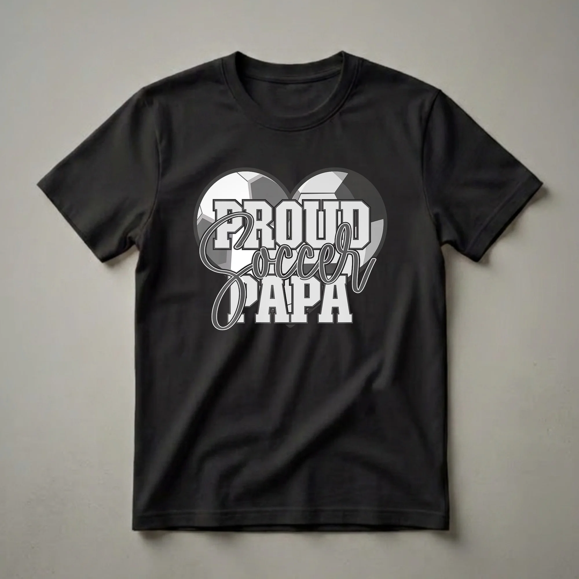 Black t-shirt with a graphic design that reads 'Proud Soccer Papa' in grey and white lettering, set against a heart shape made of two interlocking soccer balls.