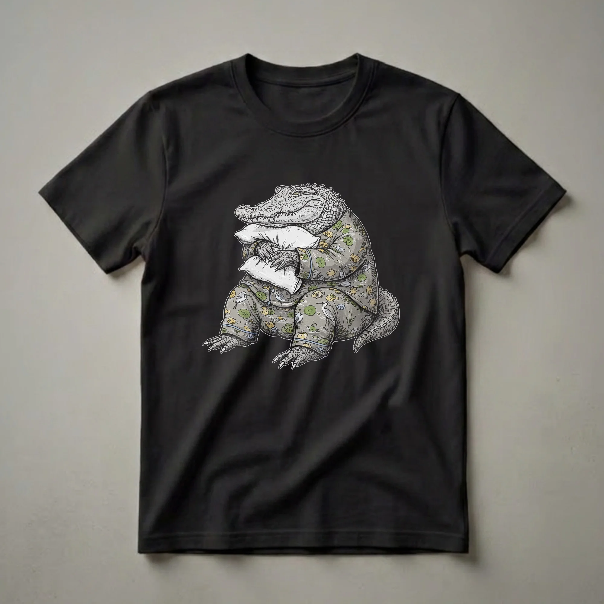 Grey graphic t-shirt featuring a cartoon alligator wearing pajamas and holding two white pillows, with a pattern of fish and lily pads on the pajamas.