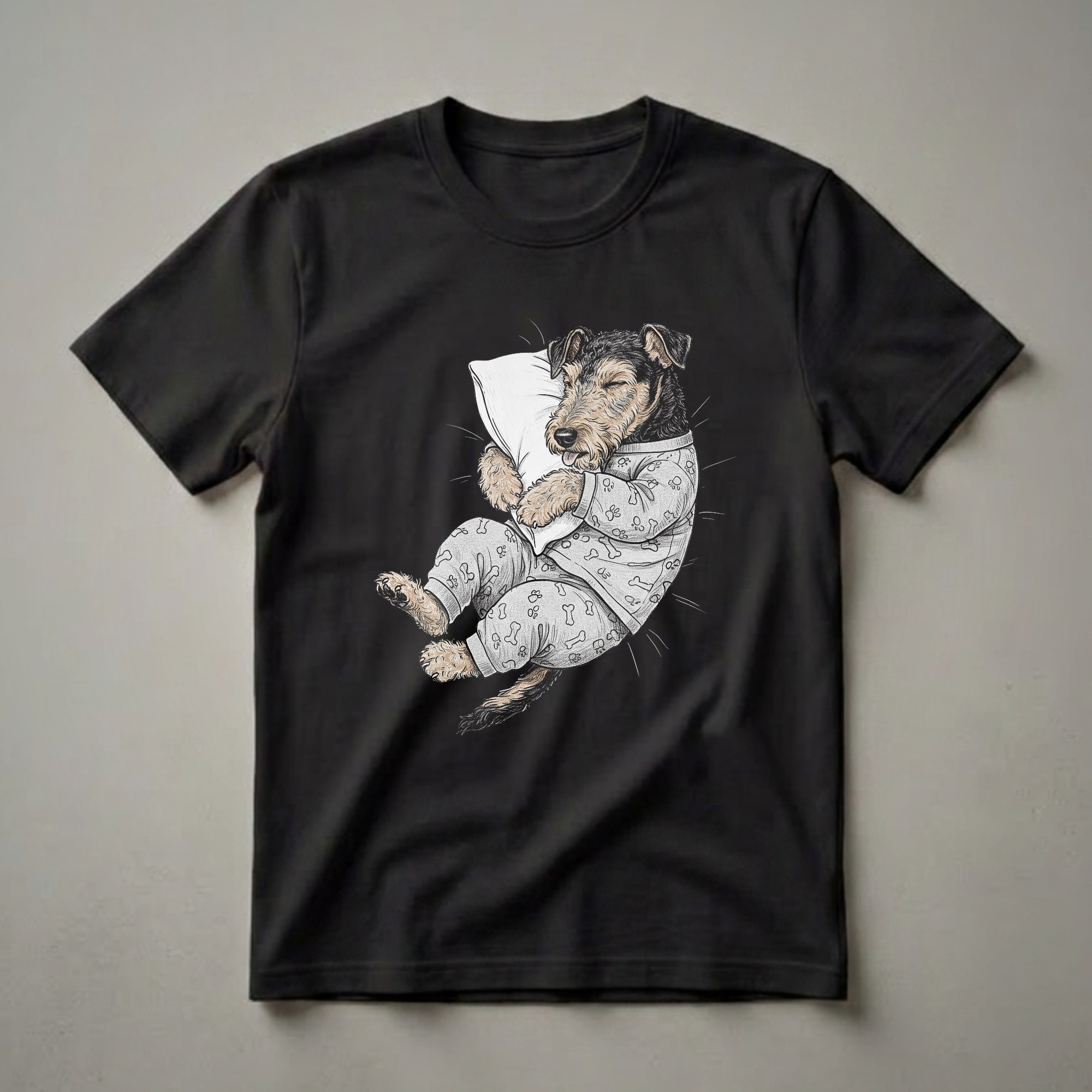 A black t-shirt featuring a whimsical illustration of a sleepy Airedale Terrier dog wearing gray pajamas with paw prints and bones, holding a white pillow.