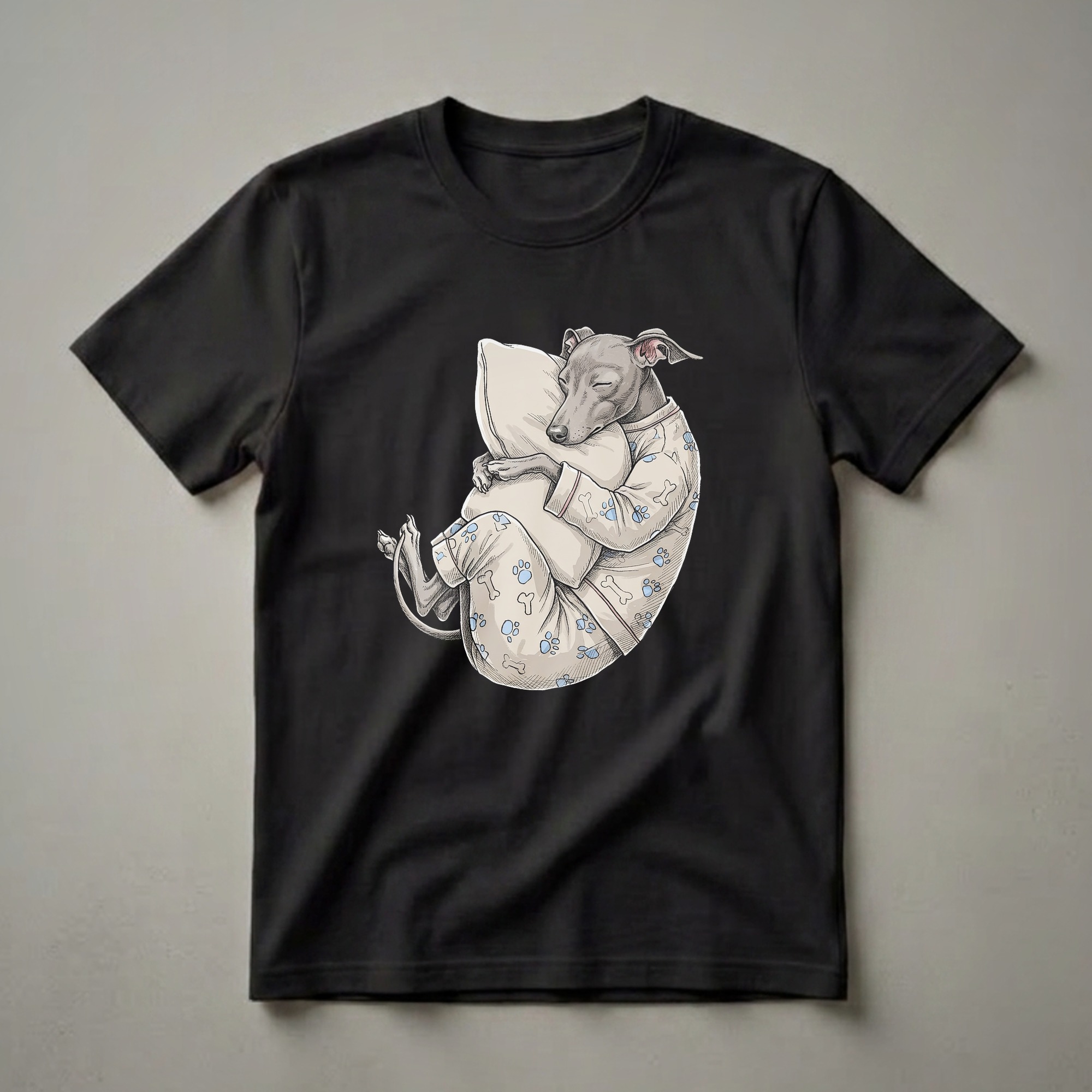 A light-colored t-shirt featuring an illustration of a grey Italian Greyhound dog sleeping peacefully while hugging a pillow, wearing pajamas with paw print and bone patterns.