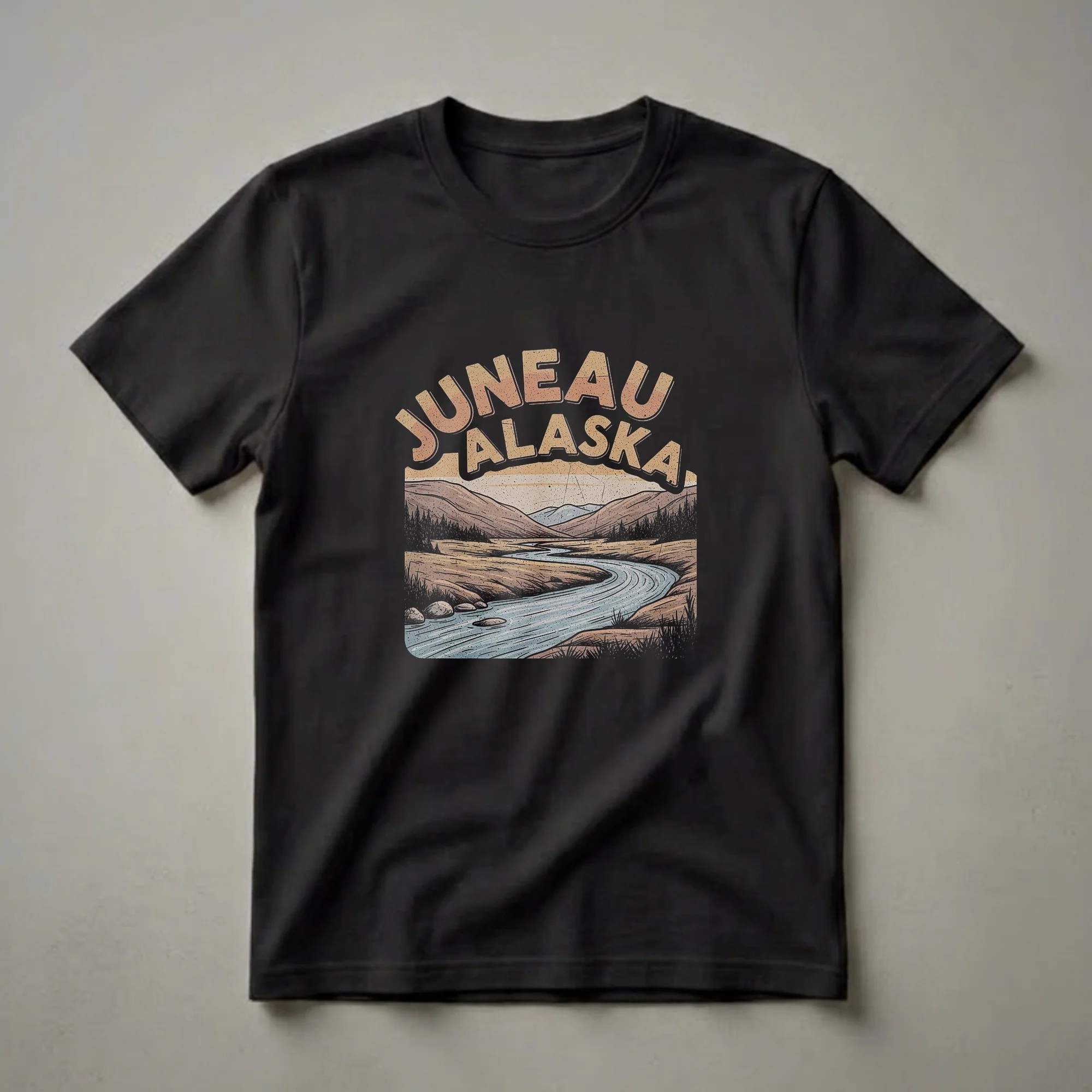 Black t-shirt with retro bold arch lettering JUNEAU ALASKA above a warm-tone illustrated river landscape with rolling hills, pine trees, and distant mountains.