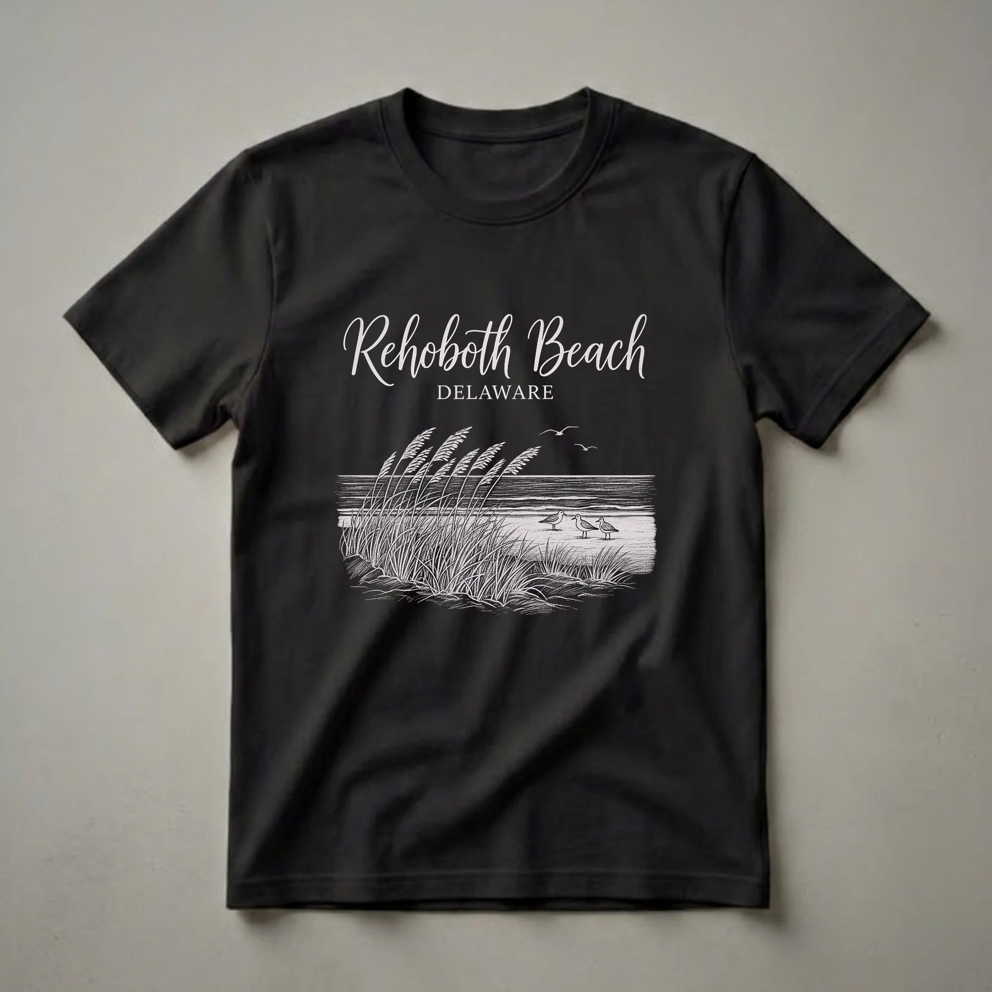 Black t-shirt with a vintage white drawing of Rehoboth Beach, Delaware, featuring sea grass, three seagulls on the sand, and ocean waves.