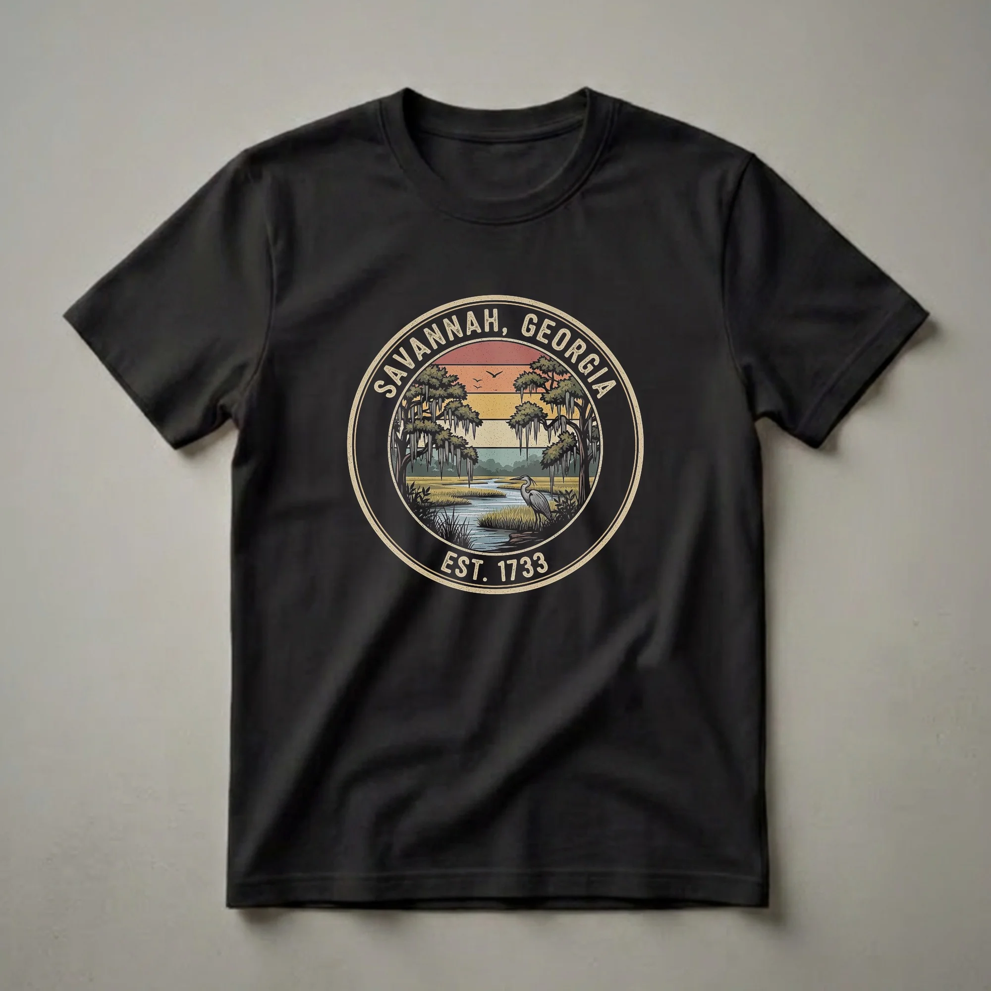 Black t-shirt with a circular retro design of a Savannah, Georgia marsh scene, featuring a heron, Spanish moss-draped trees, and a striped sunset.