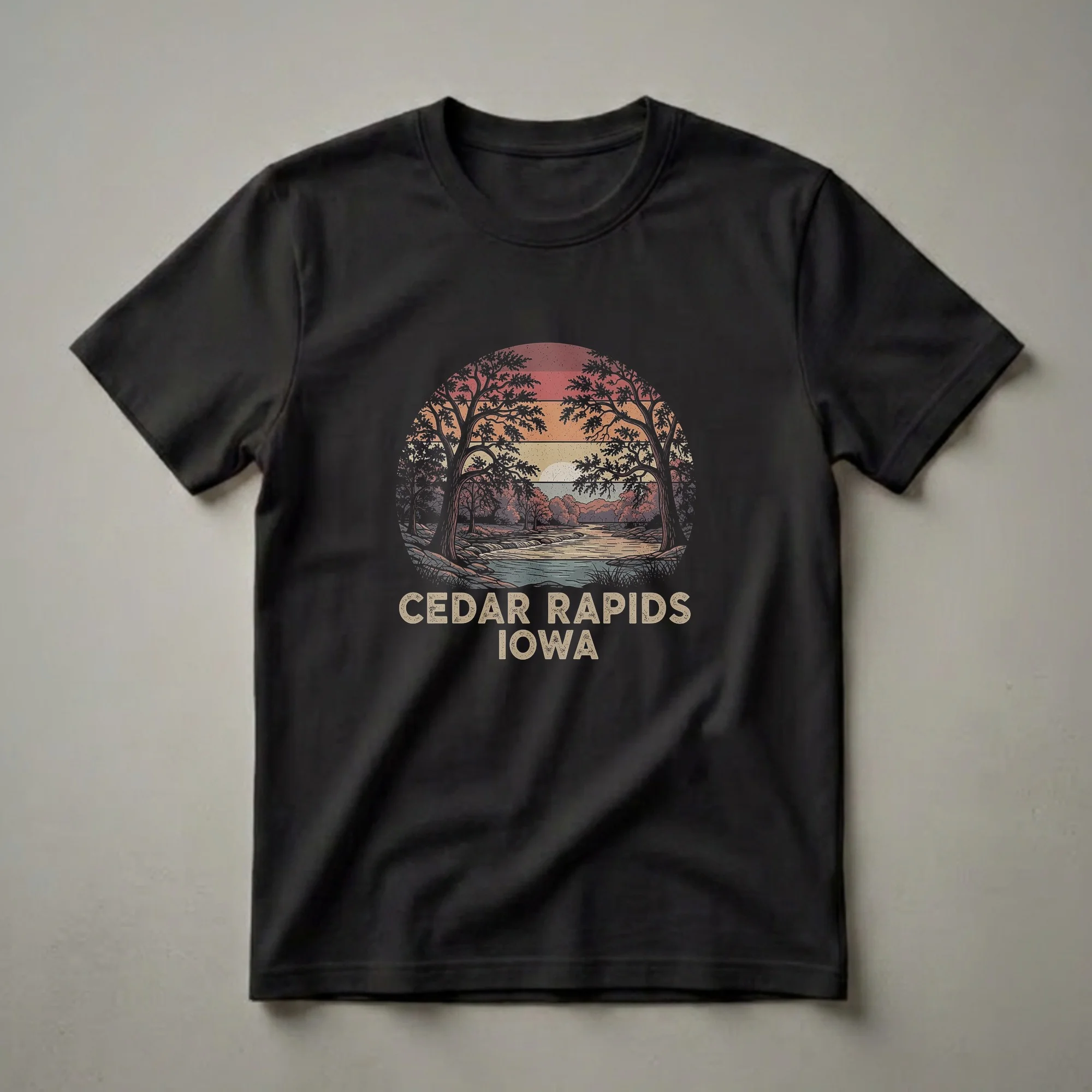Black t-shirt with a circular retro graphic of a river scene with trees and a sunset, featuring the text 'CEDAR RAPIDS IOWA' below.