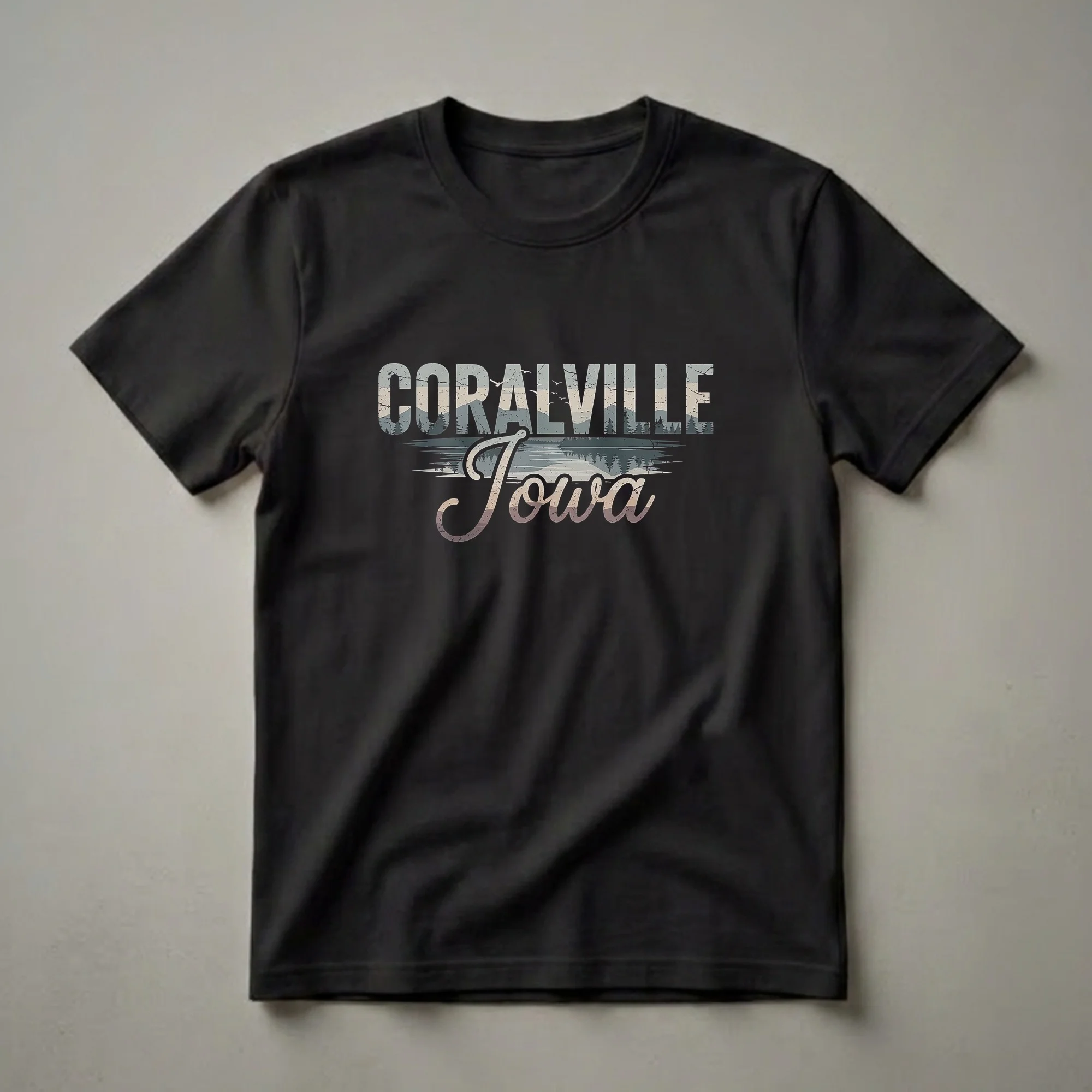 Black t-shirt with a retro graphic design featuring the words "Coralville Iowa" over a stylized landscape of hills, a lake, and pine trees.
