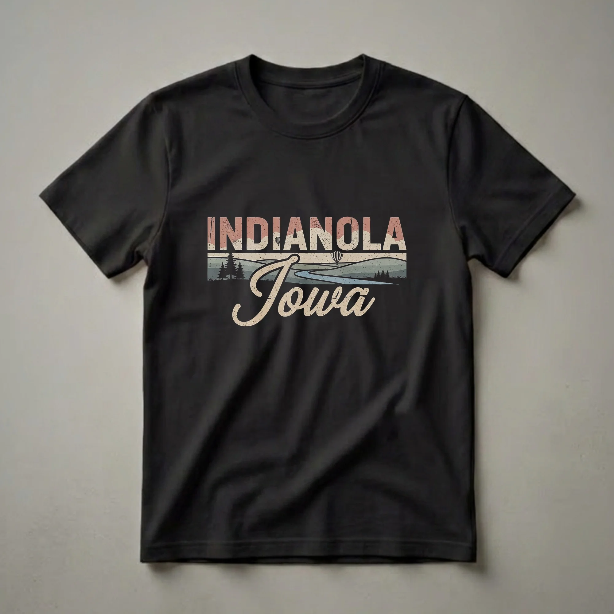 Black graphic t-shirt featuring the text "INDIANOLA Iowa" in a retro font over a stylized landscape with rolling hills, trees, a river, and hot air balloons.