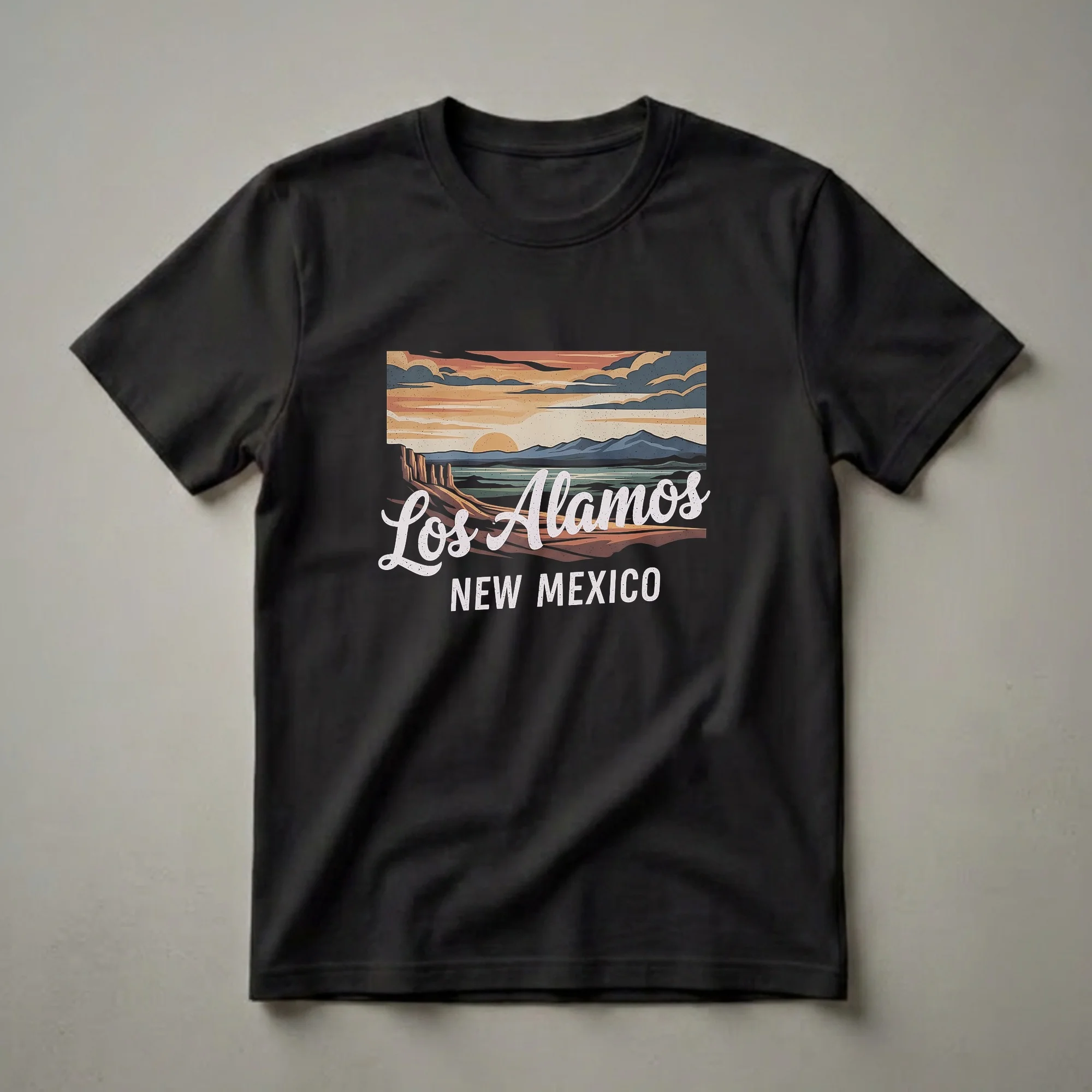 Black t-shirt with a retro graphic design depicting a sunset landscape over Los Alamos, New Mexico, featuring mesas, water, and mountains.